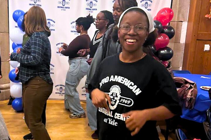 A smiling young woman wearing glasses and a Afro American Cultural Center at Yale shirt stands in the foreground at an indoor event, with other people and red, black, and white balloons in the background.