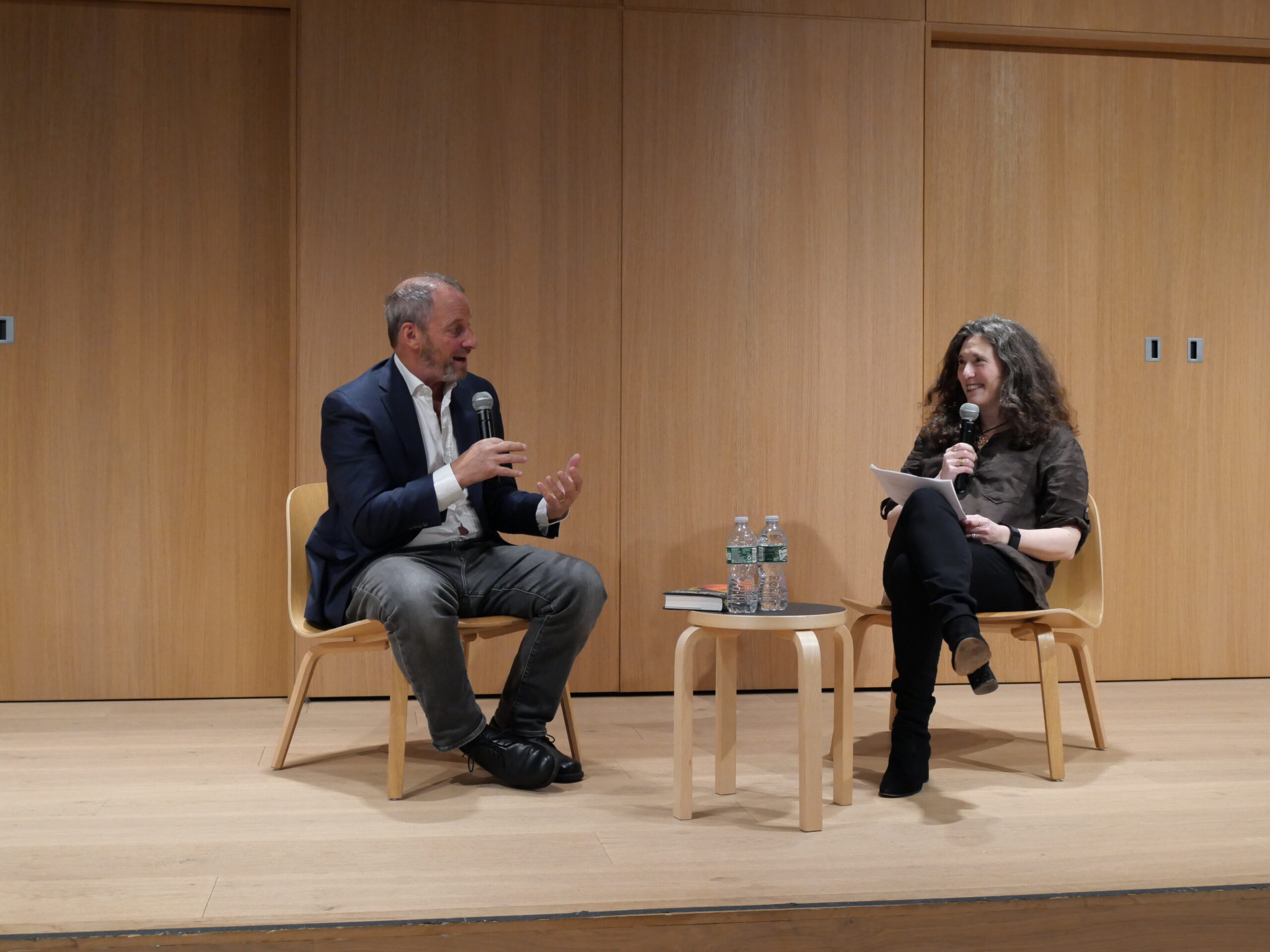 A man and a woman sit on wooden chairs on a stage, engaged in conversation with microphones. A small table with two water bottles and books is between them. Both appear to be smiling. The background is wooden paneling.