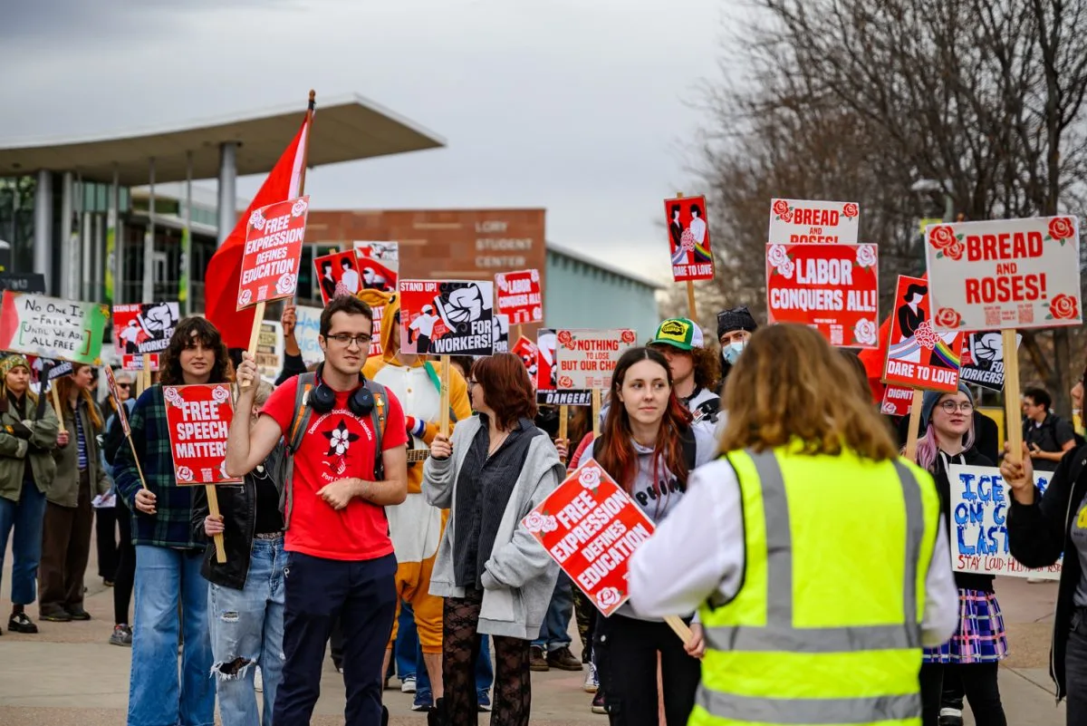 A group of protesters hold signs with messages like “Free Speech Matters” and “Bread and Roses” while marching outdoors. One person in a yellow vest stands in the foreground facing the crowd.