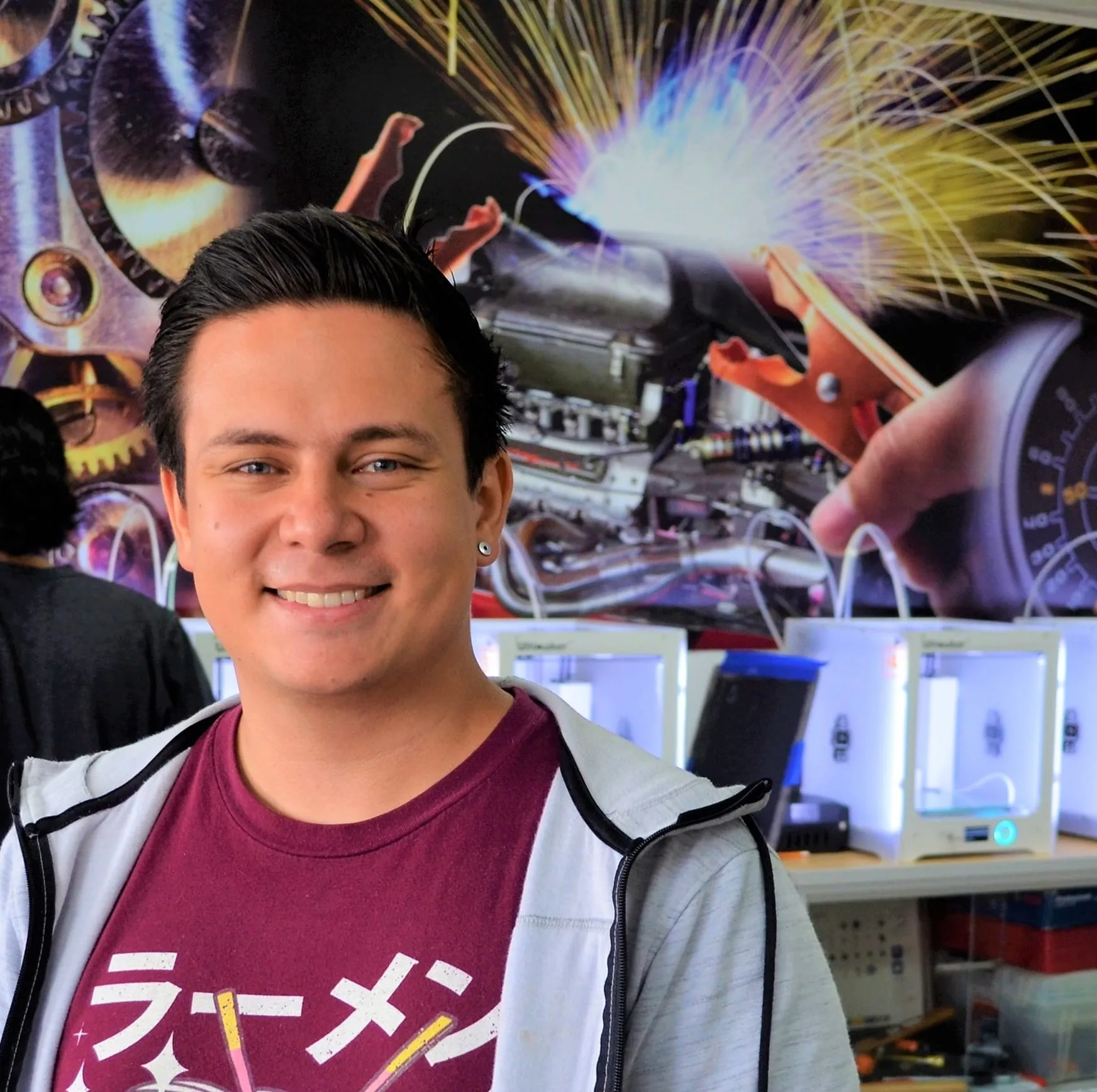 A young man smiles at the camera in a tech workshop with 3D printers and a vibrant mural of gears, sparks, and machinery in the background. He wears a maroon shirt with Japanese text and a light jacket.