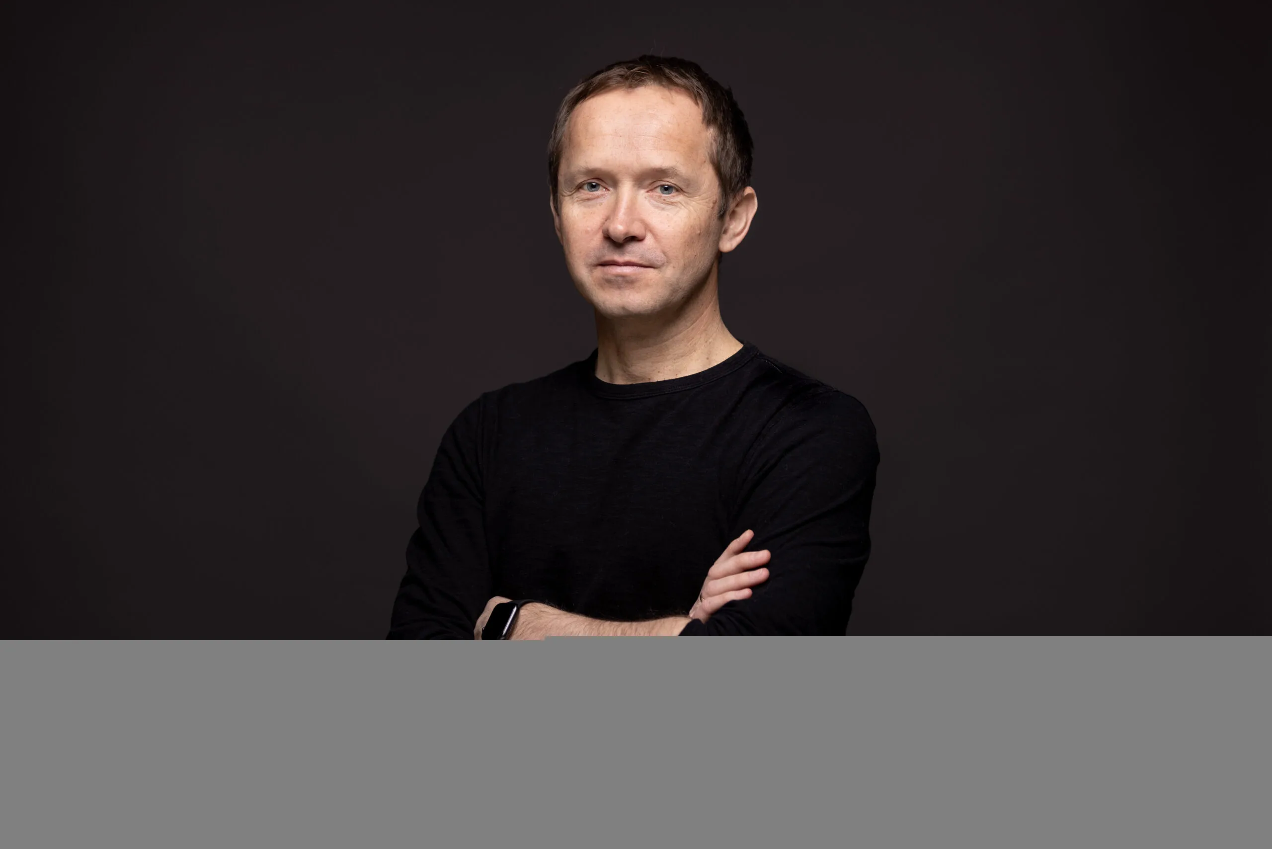 A man with short brown hair wearing a black long-sleeve shirt stands against a dark background with arms crossed, looking at the camera with a neutral expression.