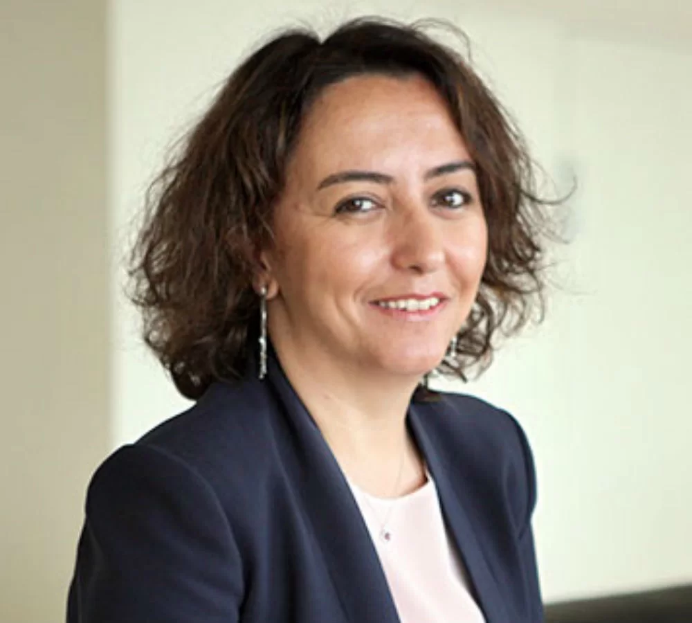 A woman with wavy brown hair wearing a navy blazer and light top smiles at the camera in a softly lit indoor setting.
