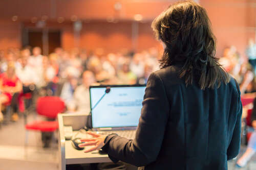 A person stands at a podium with a laptop, speaking to an audience in a conference room. The audience is out of focus, emphasizing the speakers perspective.